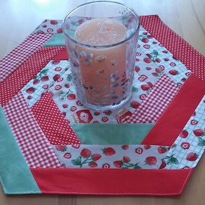 Pode incluir: Um sousplat hexagonal acolchoado com um castiçal de vidro transparente. O sousplat apresenta tecido xadrez vermelho, bolinhas vermelhas e estampa de morango. Uma vela cor de pêssego está dentro do castiçal de vidro.