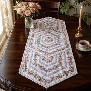 Könnte beinhalten: Ein gesteppter Tischläufer mit einem sich wiederholenden Sechseckmuster auf weißem Grund, hellvioletten Rändern und einem lebendigen Blumenmuster aus kleinen rosa, orangefarbenen und blauen Blüten, platziert auf einem dunklen Holztisch.
