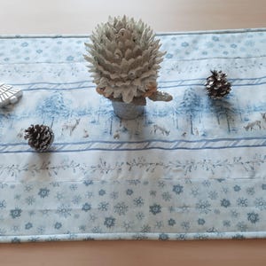 Può includere: Un runner da tavolo blu e bianco con un design a tema bosco invernale. Il runner presenta fiocchi di neve, alberi di pino e cervi. Un pigna argentata e un ornamento per albero di Natale argentato sono sul runner.