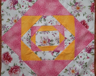 Op de afbeelding: Een roze en gele quiltblok met een bloemenpatroon. Het blok is gemaakt van vierkanten en driehoeken, en het midden is een kleiner vierkant met een bloemenpatroon.