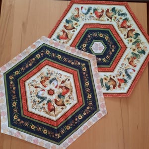 Peut inclure: Deux nappes hexagonales avec un fond crème et une bordure multicolore. Le centre de chaque nappe présente un motif circulaire avec des coqs et des accents floraux.