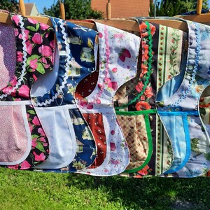 May include: A row of six fabric aprons hanging on wooden hangers, each featuring distinct patterns including floral, strawberry, and striped designs. The aprons are finished with decorative rickrack trim and solid-colored pockets with white piping.