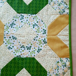May include: A square quilted fabric mat featuring a geometric patchwork design. The pattern includes sections of white fabric with green and yellow butterflies, solid mustard yellow triangles, and cream fabric with small green dots, all bordered by green trim.