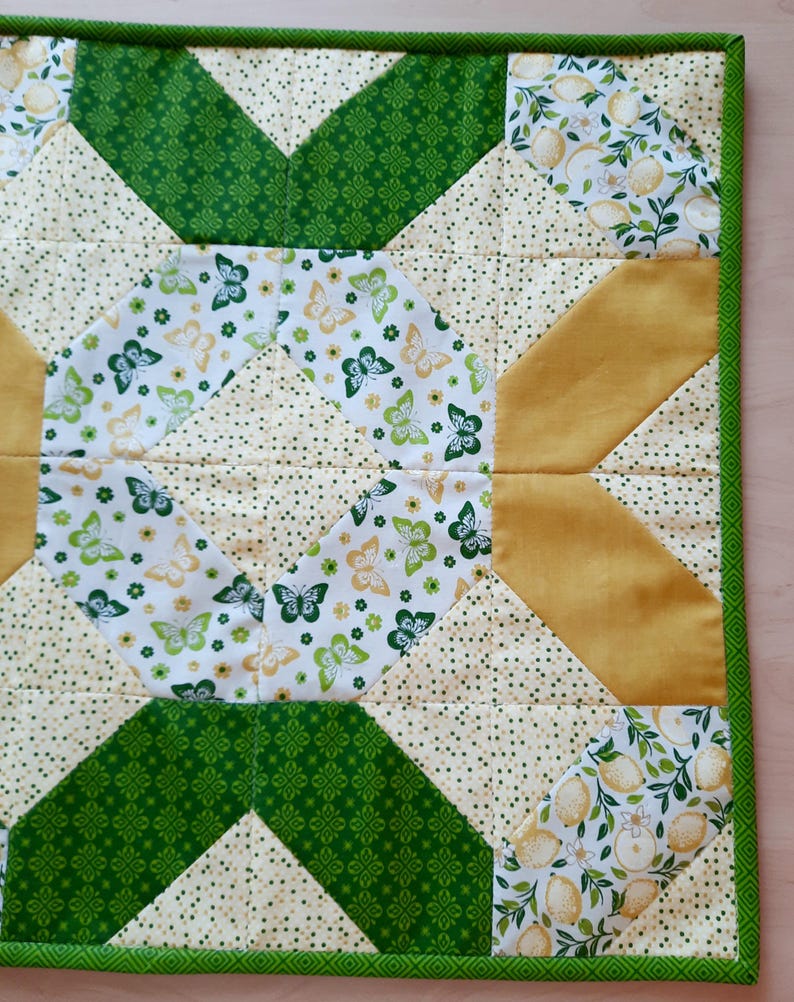 May include: A square quilted fabric mat featuring a geometric patchwork design. The pattern includes sections of white fabric with green and yellow butterflies, solid mustard yellow triangles, and cream fabric with small green dots, all bordered by green trim.
