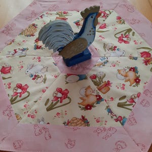 Op de afbeelding: Een houten haanfiguur in blauw en bruin staat op een zeshoekige tafelloper. De loper heeft een lichtgele midden met een bloemen- en dierenprint en een roze rand met een herhalend patroon.