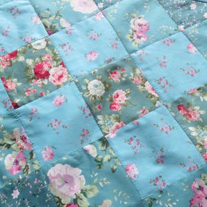 Op de afbeelding: Een gewatteerde tafelloper met een patchwork ontwerp. De vierkanten zijn lichtblauw en hebben bloempatronen in roze, rood en wit. De loper is afgewerkt met een bijpassende blauwe stof.