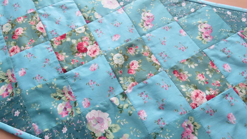 Op de afbeelding: Een gewatteerde tafelloper met een patchwork ontwerp. De vierkanten zijn lichtblauw en hebben bloempatronen in roze, rood en wit. De loper is afgewerkt met een bijpassende blauwe stof.