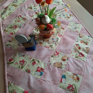 Peut inclure: Chemin de table patchwork rose et vert avec des motifs floraux et de Pâques. Un petit pot en céramique avec des œufs et une figurine de poulet est posé sur le chemin de table, ainsi qu'un coq en bois et des tulipes oranges.