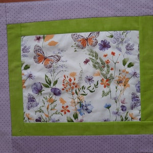 May include: A white fabric with a green border and a purple border with white polka dots. The fabric has a floral pattern with butterflies and flowers in shades of pink, purple, orange, and green.