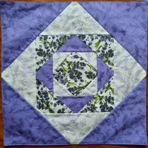 Könnte beinhalten: Eine lila und weiße Patchwork-Decke mit geometrischem Muster, das mit Lavendelblüten verziert ist. Die Decke besteht aus verschiedenen Stoffen mit unterschiedlichen Texturen und Mustern.