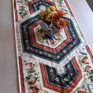 Può includere: Runner da tavolo trapuntato con un motivo di galli e girasoli. Il runner presenta uno sfondo color crema con disegni geometrici rossi, blu navy e verdi. Una statuetta di gallo in legno e girasoli in vaso sono posti sopra.