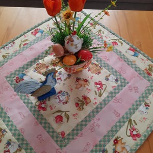 Könnte beinhalten: Dekorativer Tischläufer zum Thema Ostern mit Blumen- und Hasenmuster. Der Läufer hat ein zentrales Quadrat mit einer Hahnfigur und einem Korb mit Eiern. Orangefarbene Tulpen und ein Keramik-Huhn befinden sich in der Mitte.