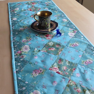 Könnte beinhalten: Türkisfarbener, gesteppter Tischläufer mit floralem Muster. Eine dekorative Tasse und Untertasse mit Goldrand liegen auf dem Läufer. Eine kleine blaue Figur befindet sich ebenfalls auf dem Läufer.