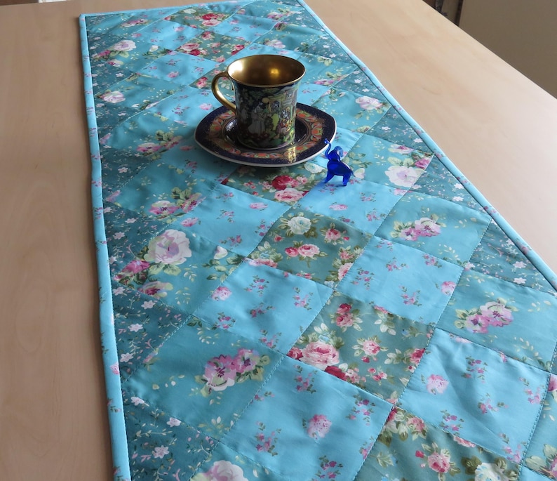 Op de afbeelding: Een turquoise gewatteerde tafelloper met een bloemenpatroon. Een decoratieve kop en schotel met een gouden rand staan op de loper. Een klein blauw beeldje staat ook op de loper.