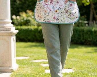 May include: A half apron featuring a white fabric with a multicolored floral print, accented by solid teal side panels and teal trim. The apron includes two front pockets and is worn over a gray t-shirt and sage green trousers.