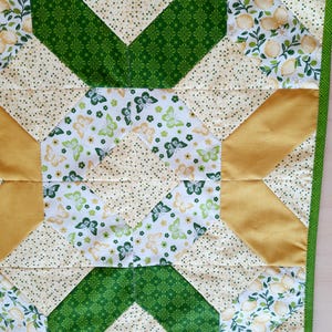 May include: A square quilted fabric piece featuring a geometric star pattern. The design incorporates patches of white fabric with yellow lemon prints, green butterfly motifs, solid mustard yellow triangles, and cream fabric with small green dots.