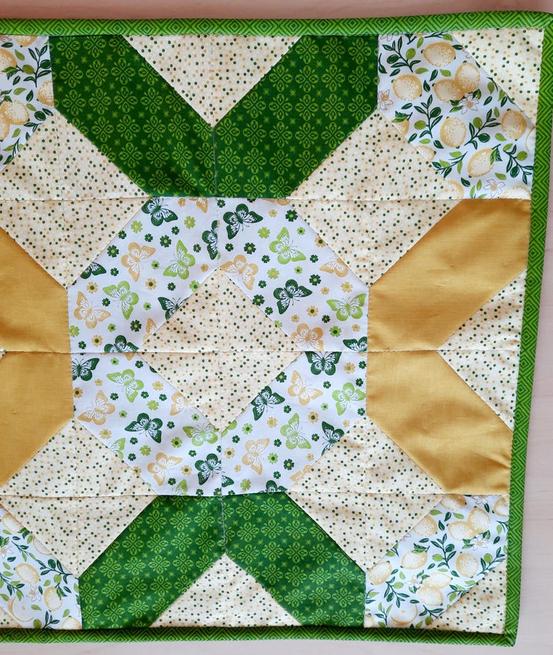 May include: A square quilted fabric piece featuring a geometric star pattern. The design incorporates patches of white fabric with yellow lemon prints, green butterfly motifs, solid mustard yellow triangles, and cream fabric with small green dots.