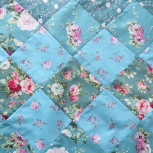 Op de afbeelding: Een gewatteerde stof met een ruitpatroon. Het ontwerp toont afwisselende vierkanten van lichtblauw en bloemenstof. De bloemenstof heeft roze en witte rozen op een teal achtergrond. De quilt wordt waarschijnlijk gebruikt voor beddengoed of woondecoratie.