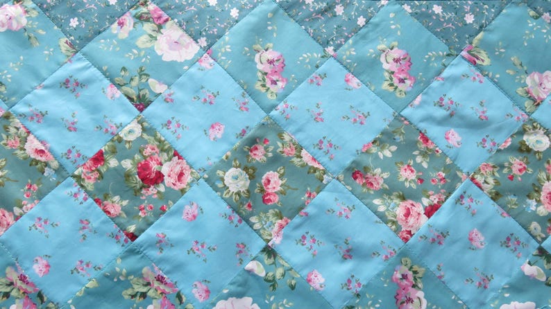 Op de afbeelding: Een gewatteerde stof met een ruitpatroon. Het ontwerp toont afwisselende vierkanten van lichtblauw en bloemenstof. De bloemenstof heeft roze en witte rozen op een teal achtergrond. De quilt wordt waarschijnlijk gebruikt voor beddengoed of woondecoratie.