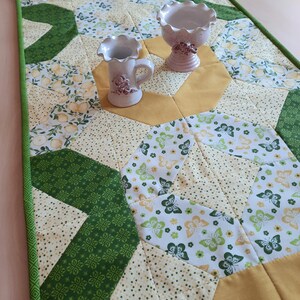 Puede incluir: Un camino de mesa acolchado hecho a mano con un borde verde. El camino de mesa presenta un diseño de patchwork con telas amarillas, verdes y blancas. El patrón incluye motivos florales y de mariposas. Dos pequeños jarrones decorativos de cerámica se encuentran sobre el camino de mesa.