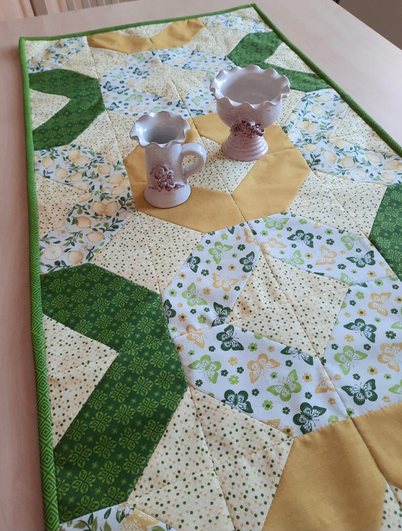 May include: A rectangular quilted table runner featuring a patchwork design of green and yellow fabrics with patterns of butterflies, lemons, and small dots. Two small, light-colored ceramic vessels with ruffled edges sit on the runner.