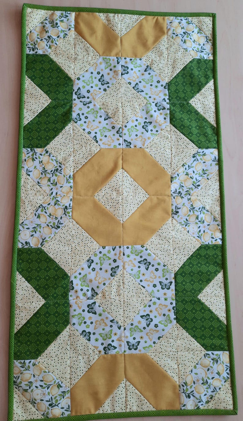May include: A rectangular quilted table runner featuring a patchwork design with green, yellow, and white fabric. The pattern includes sections with lemon prints, butterfly motifs, and solid mustard yellow diamonds, all bordered by a green trim.