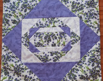 Op de afbeelding: Een paars-witte quiltblok met een geometrisch ontwerp met een lavendelbloemprint. De blok is gemaakt van vierkanten en driehoeken, waardoor een spiraalpatroon ontstaat.