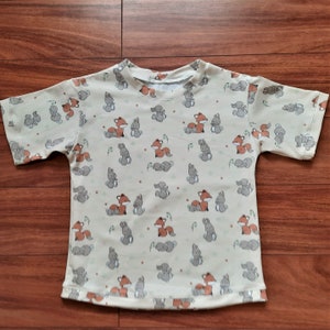 Puede incluir: Una camiseta beige de manga corta con un patrón repetido de zorros marrones y conejos grises. Los zorros se toman de la mano y los conejos están sentados en posición vertical.