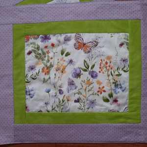 May include: A lavender and green fabric placemat with a floral and butterfly design. The placemat has a white background with a repeating pattern of butterflies, flowers, and leaves.