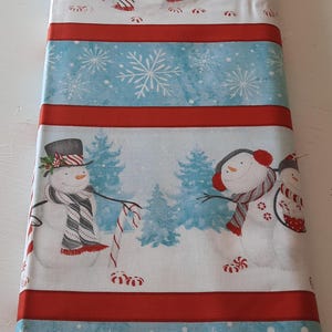 May include: Red and white striped fabric with a blue background featuring a winter scene with snowmen and candy canes. The fabric has a repeating pattern of snowflakes.