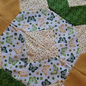 May include: A quilted fabric piece featuring a geometric pattern with alternating sections of solid mustard yellow, green fabric with a white clover print, and a white background printed with small yellow flowers and green and yellow butterflies.