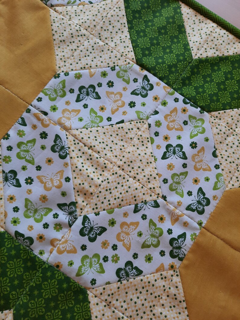 May include: A quilted fabric piece featuring a geometric pattern with alternating sections of solid mustard yellow, green fabric with a white clover print, and a white background printed with small yellow flowers and green and yellow butterflies.