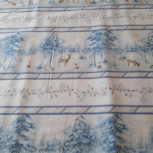 Può includere: Un tessuto bianco con un motivo ripetuto di alberi blu, cervi e conigli in una foresta invernale innevata. Il tessuto ha un bordo a strisce blu e bianco.