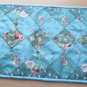 Op de afbeelding: Een gewatteerde tafelloper met bloemenpatroon. De loper heeft een patchwork-ontwerp van turquoise en bloemenstof vierkanten. De bloemenstof heeft roze en witte rozen. De randen zijn afgewerkt met een turquoise rand. Ongeveer 100 cm lang.