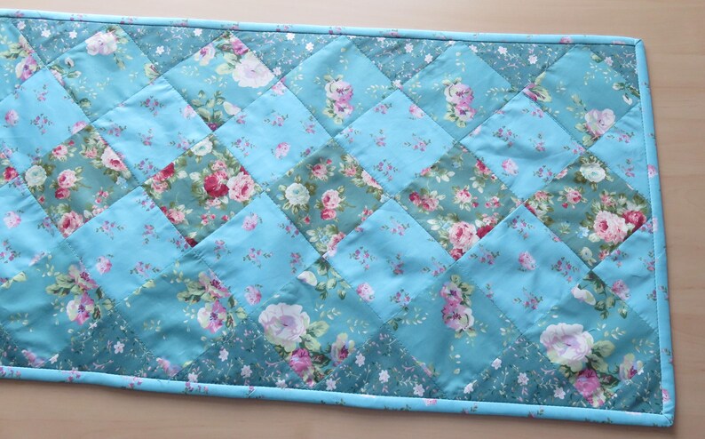 Op de afbeelding: Een gewatteerde tafelloper met bloemenpatroon. De loper heeft een patchwork-ontwerp van turquoise en bloemenstof vierkanten. De bloemenstof heeft roze en witte rozen. De randen zijn afgewerkt met een turquoise rand. Ongeveer 100 cm lang.
