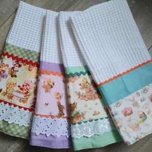 May include: Four white waffle weave kitchen towels with decorative patterned bottoms. The towels feature colorful designs with characters, bunnies, and Easter eggs. Each towel has a different color scheme, including green, purple, and blue.