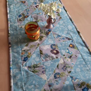 Op de afbeelding: Een blauwe en witte gestikte tafelrunner met een bloemenpatroon. De runner is versierd met een kleine vaas met witte bloemen en een glazen kaarsenhouder.