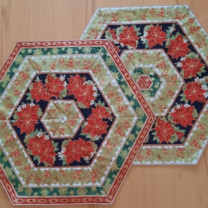Può includere: Due tovagliette esagonali trapuntate a tema natalizio. Le tovagliette presentano un disegno centrale di stelle di Natale rosse su sfondo blu navy, circondate da bordi verdi e dorati. I bordi sono rifiniti con un bordo rosso e bianco.