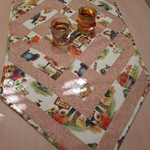 Peut inclure: Chemin de table matelassé sur le thème de la récolte d'automne. Le chemin de table présente un motif patchwork avec des images de citrouilles, d'épouvantails et de tournesols. Le fond est un mélange de blanc et de marron, avec un motif quadrillé. Deux verres sont posés dessus.