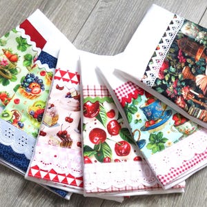 Può includere: Una collezione di strofinacci da cucina decorativi con vari disegni di frutta e fiori. Ogni strofinaccio presenta un motivo unico, tra cui fragole, ciliegie e composizioni floreali, con bordi in pizzo decorativi. Gli strofinacci sono disposti su una superficie di legno.