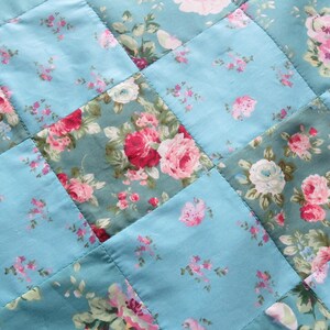 Op de afbeelding: Close-up van een gewatteerde stof met een bloemenpatroon. De quilt heeft een patchwork-ontwerp met afwisselende vierkanten van effen lichtblauwe en bloemenprintstof. De bloemenprint bevat roze en witte rozen.