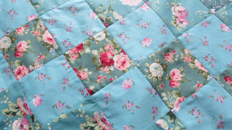Op de afbeelding: Close-up van een gewatteerde stof met een bloemenpatroon. De quilt heeft een patchwork-ontwerp met afwisselende vierkanten van effen lichtblauwe en bloemenprintstof. De bloemenprint bevat roze en witte rozen.