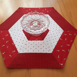 Peut inclure: Chemin de table hexagonal avec un motif patchwork rouge et blanc. Le chemin de table présente une bordure en vichy rouge, des sections blanches avec de minuscules cœurs rouges et des accents floraux rouges. Un bol en verre transparent est au centre.