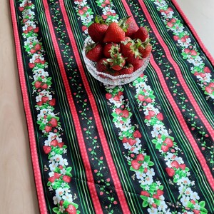 Könnte beinhalten: Ein rechteckiger Tischläufer mit schwarzem Hintergrund, roten Streifen und einem Erdbeer- und Blumenmuster. Eine klare Glasschale mit frischen Erdbeeren steht auf dem Läufer. Der Läufer ist aus Stoff.