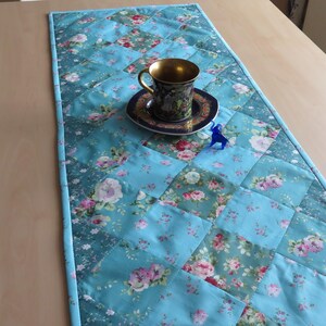 Op de afbeelding: Een gewatteerde tafelloper in tinten turquoise, met een patchwork ontwerp met bloempatronen in roze en wit. Een decoratieve theekop en schotel met gouden rand staan op de loper, samen met een klein blauw beeldje. De loper heeft een lichtblauwe rand.