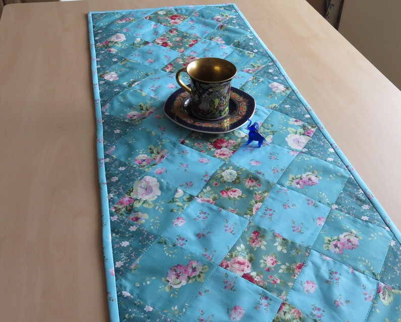 Op de afbeelding: Een gewatteerde tafelloper in tinten turquoise, met een patchwork ontwerp met bloempatronen in roze en wit. Een decoratieve theekop en schotel met gouden rand staan op de loper, samen met een klein blauw beeldje. De loper heeft een lichtblauwe rand.