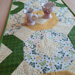 May include: A rectangular quilted table runner featuring a patchwork design of green, yellow, and white fabrics with butterfly and floral patterns. Two white ceramic vessels with ruffled rims and small floral accents sit on the runner.
