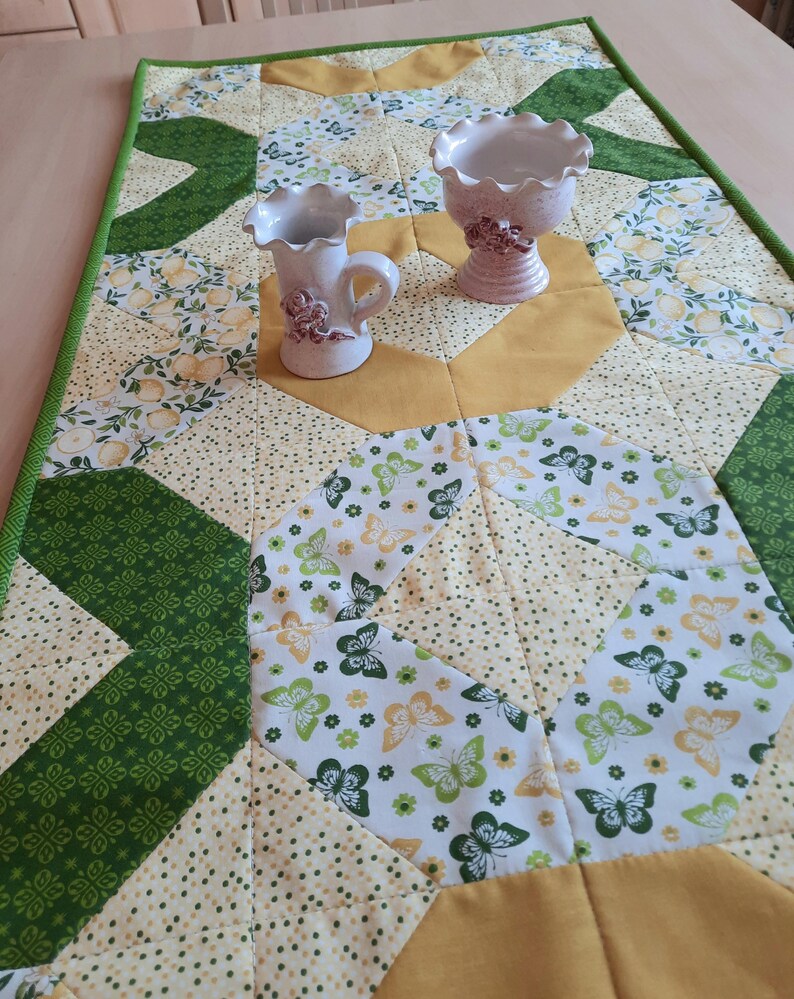 May include: A rectangular quilted table runner featuring a patchwork design of green, yellow, and white fabrics with butterfly and floral patterns. Two white ceramic vessels with ruffled rims and small floral accents sit on the runner.