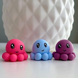 May include: Three colorful octopus figurines. The octopus on the left is pink with glitter, the middle one is light blue, and the right one is purple. Each has a smiling face and eight tentacles. A white textured background is behind them.