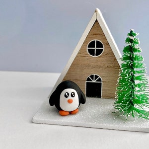 May include: A small wooden cabin with a white roof and a green pine tree. A black and white penguin figurine stands in front of the cabin. The scene is decorated with white glitter to resemble snow.